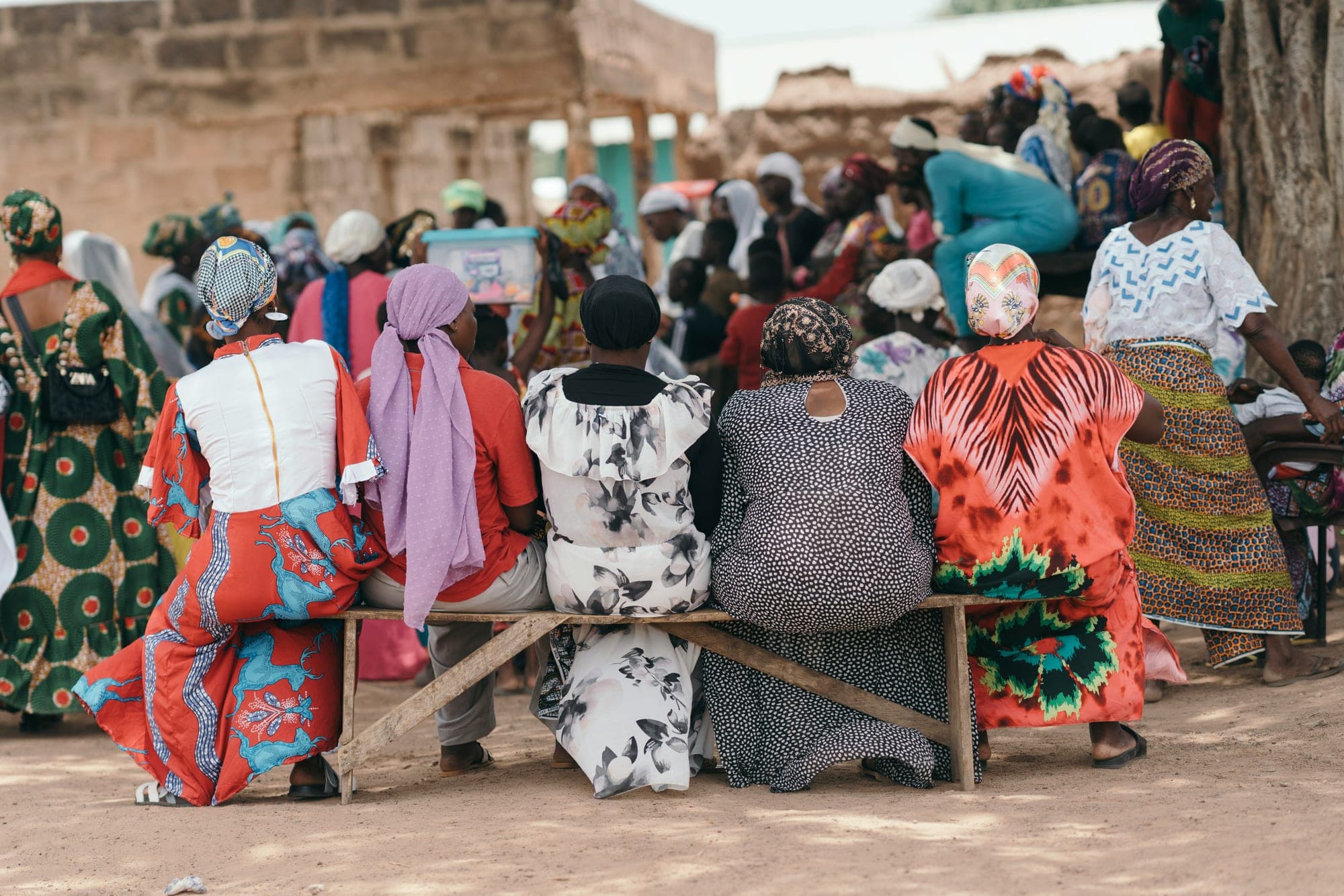 Women in colourful dress seated at a community gathering, a crowd behind them